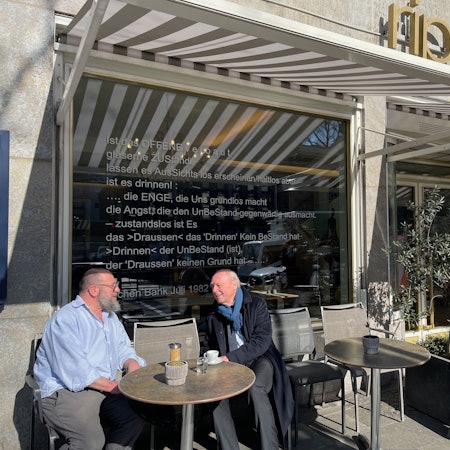 Ein Mann mit Bart und ein Senior in Schal und Mantel sitzen an einem Tisch draußen vor einem Restaurant.
