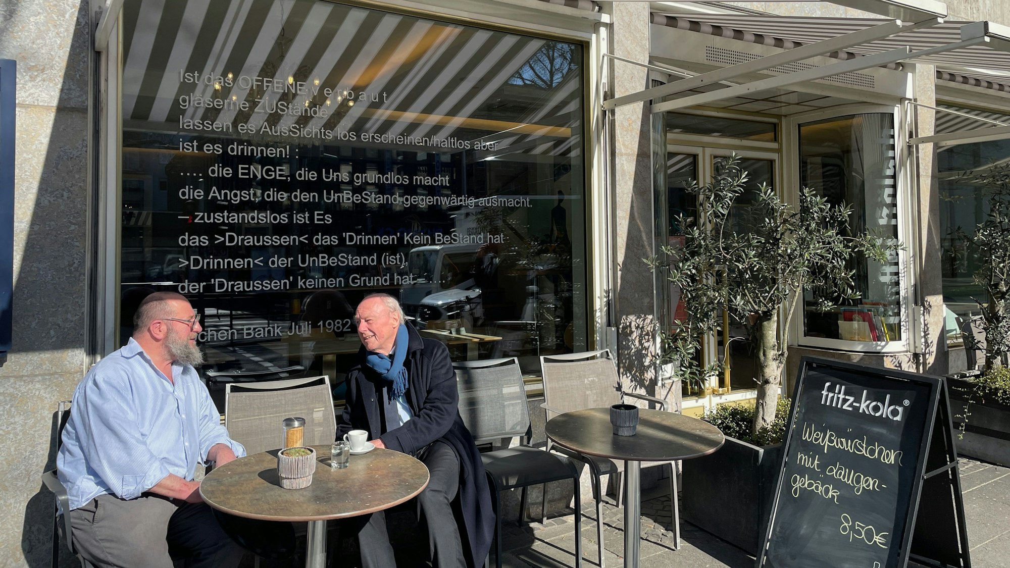 Ein Mann mit Bart und ein Senior in Schal und Mantel sitzen an einem Tisch draußen vor einem Restaurant.