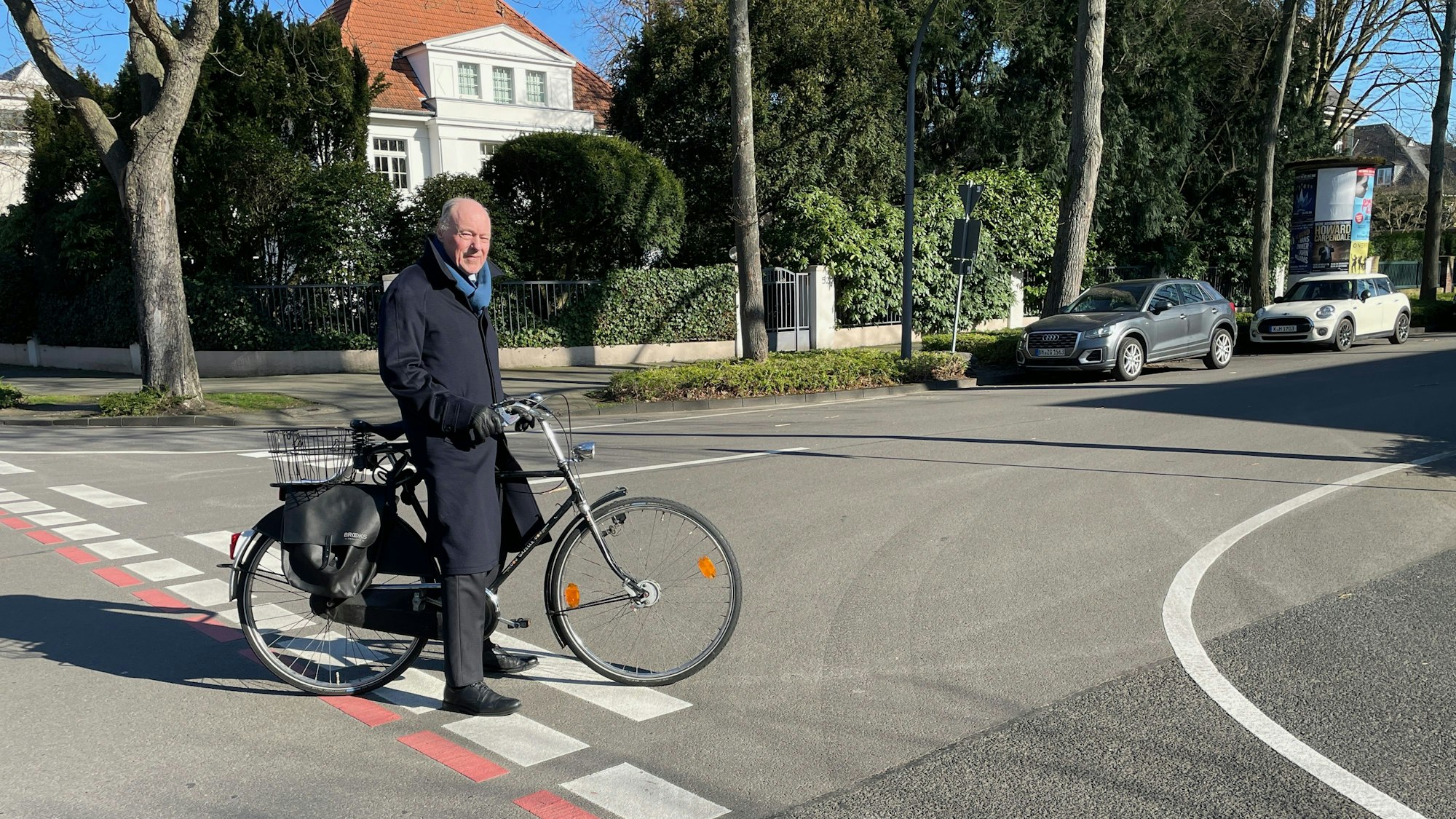 Ein Mann mit einem Hollandrad steht auf einer Straßenkreuzung, im Hintergrund ist eine stattliche Villa zu sehen.