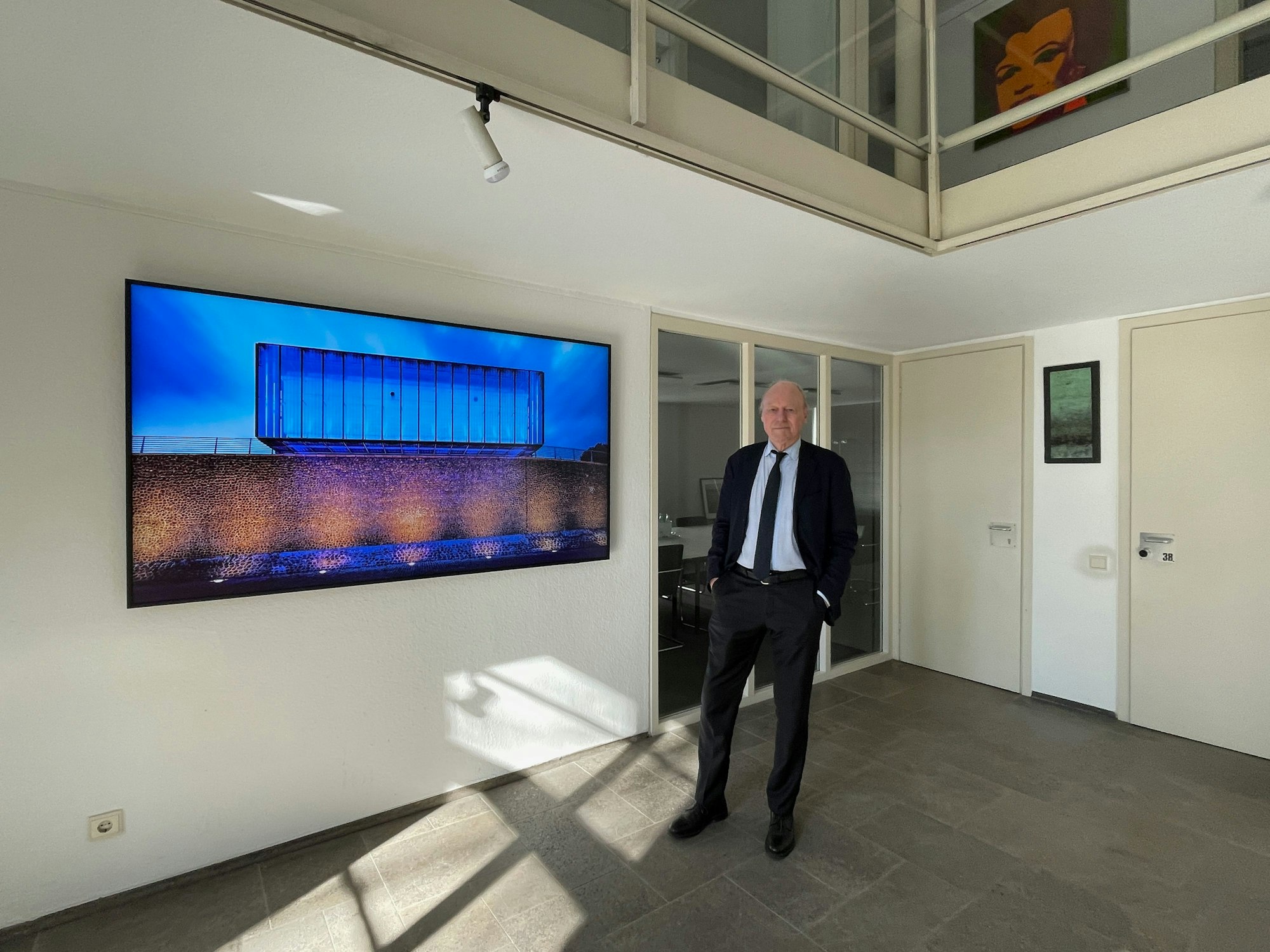 Ein Mann im Anzug steht in einem Raum, an der Wand hängt ein großformatiges Bild vom Hochwasserpumpwerk in Bayenthal.