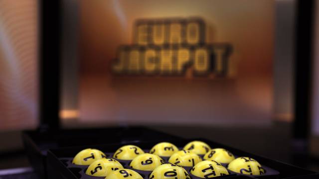 Das Bild zeigt mehrere gelbe Lotteriekugeln mit schwarzen Nummern, die ordentlich in einem schwarzen Tablett angeordnet sind. Im unscharfen Hintergrund ist das beleuchtete Eurojackpot-Logo zu erkennen.