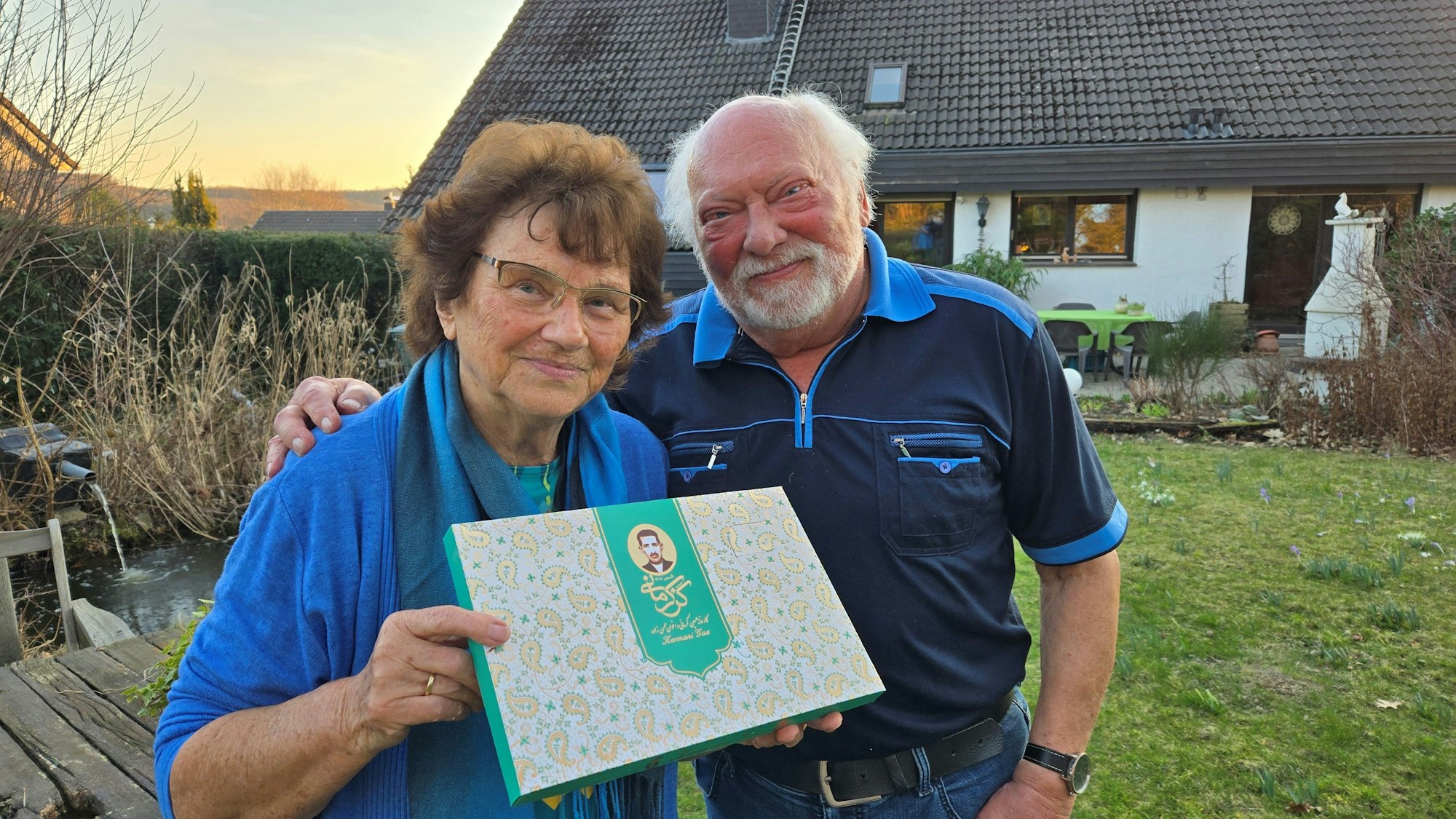 Ein älteres Ehepaar steht vor seinem Haus, sie hält eine Schachtel.