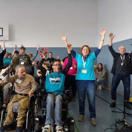 Menschen mit und ohne Behinderung werfen die Arme nach oben.