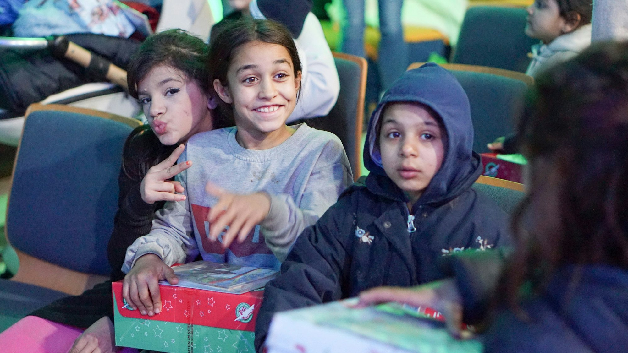 Drei Kinder sitzen auf Stühlen. Sie haben Geschenkekartons auf dem Schoß.