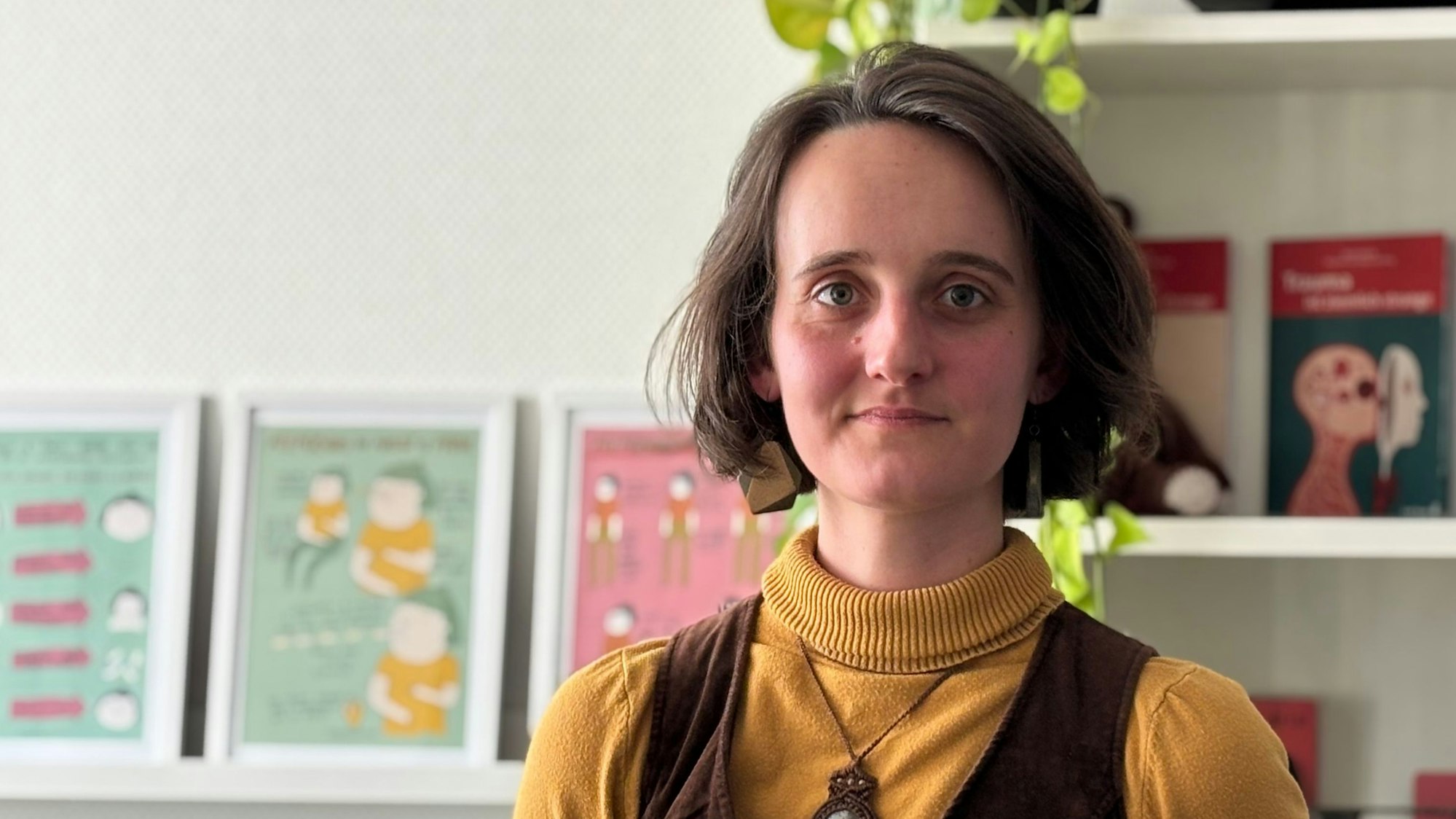 Eine Frau steht vor einem Büroschrank. Sie trägt kinnlanges Haar, einen gelben Rollkragenpullover und eine braune Weste.