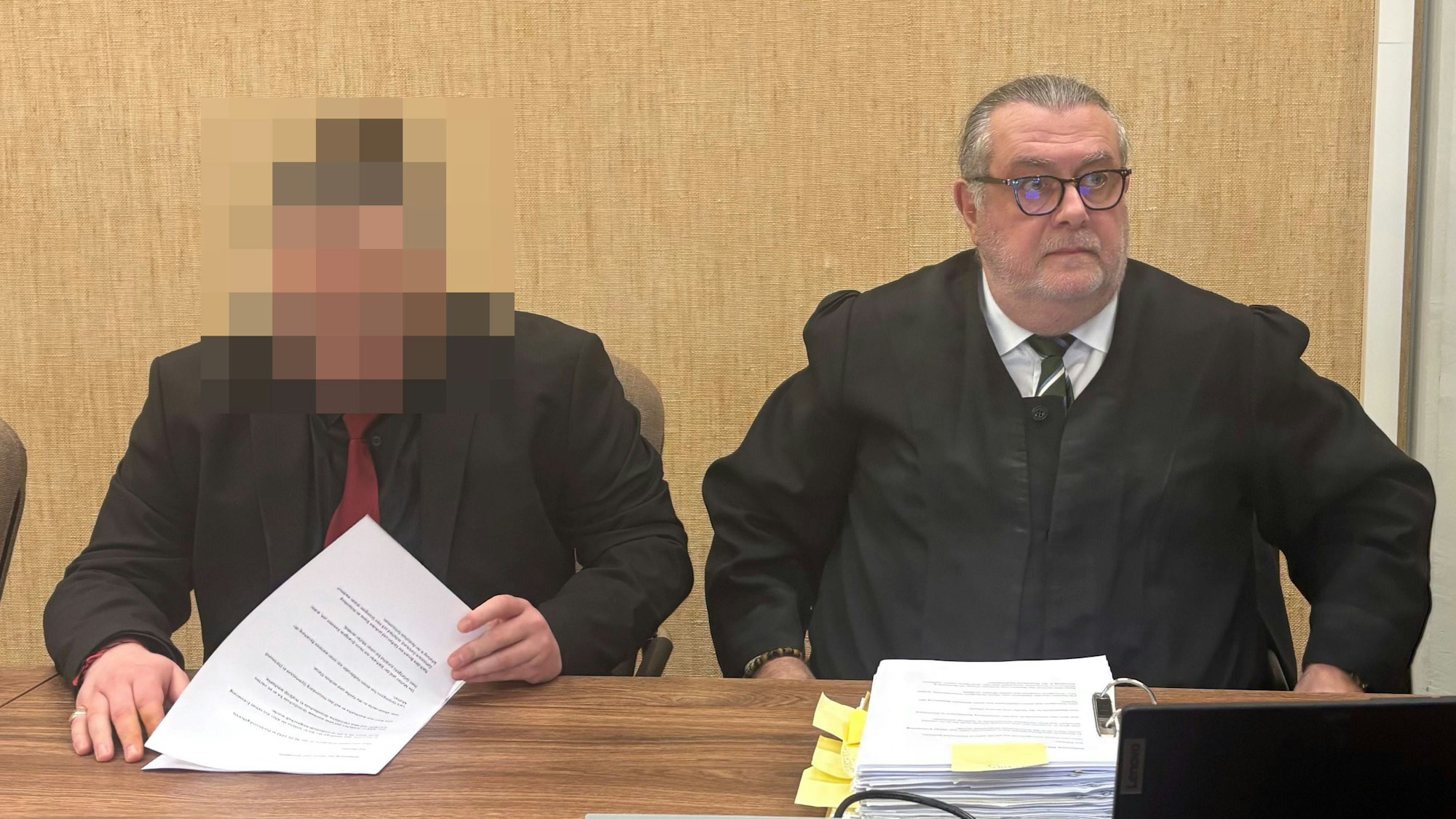 Der Angeklagte sitzt im schwarzen Anzug mit roter Krawatte neben seinem Verteidiger Thomas Düber.