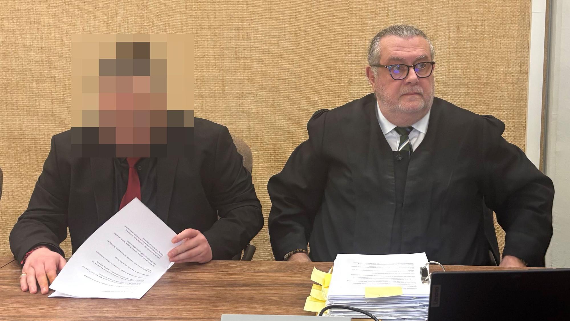 Der Angeklagte trägt einen schwarzen Anzug und eine rote Krawatte.