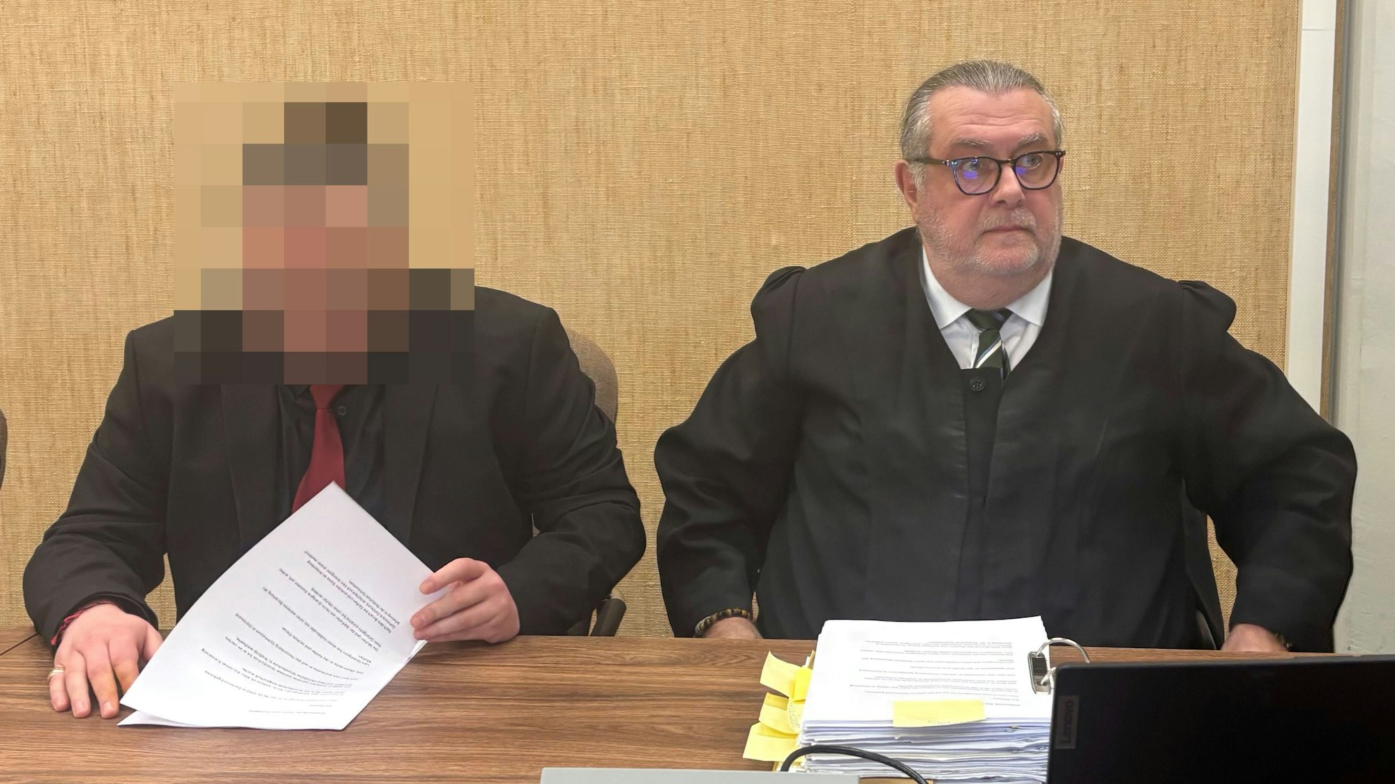 Der Angeklagte trägt einen schwarzen Anzug und eine rote Krawatte.