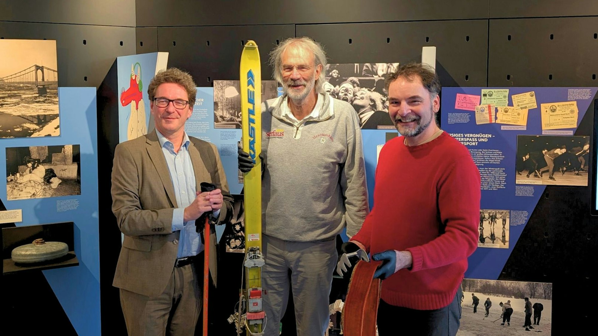 Drei Männer stehen vor einer Bilderwand, der Mann in der Mitte hält ein Paar Skier.
