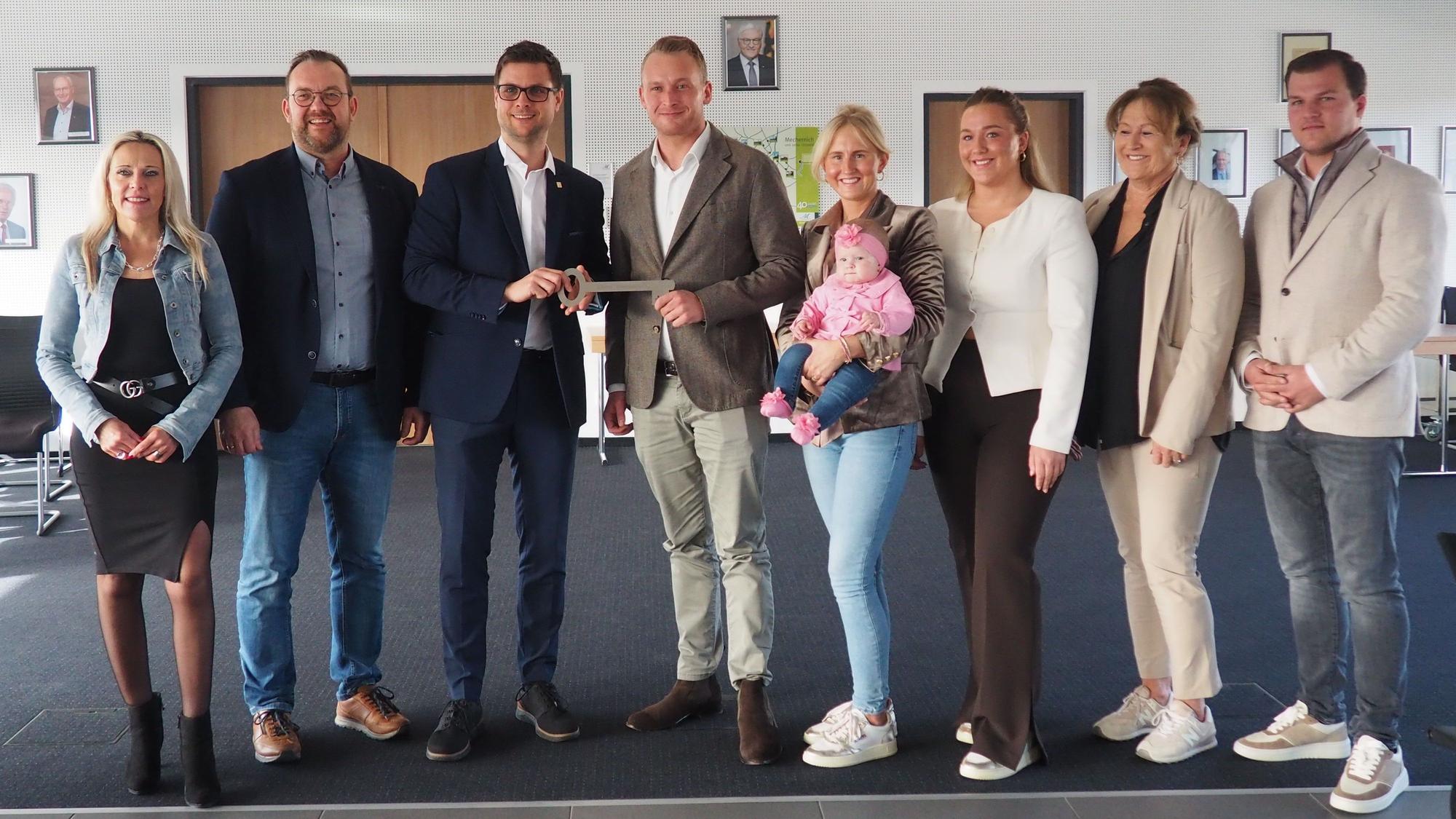 Acht Männer und Frauen stehen in einer Reihe. Bürgermeister Fingel übergibt Roland Barth einen großen Schlüssel. Eine Frau hält ein Baby im Arm.