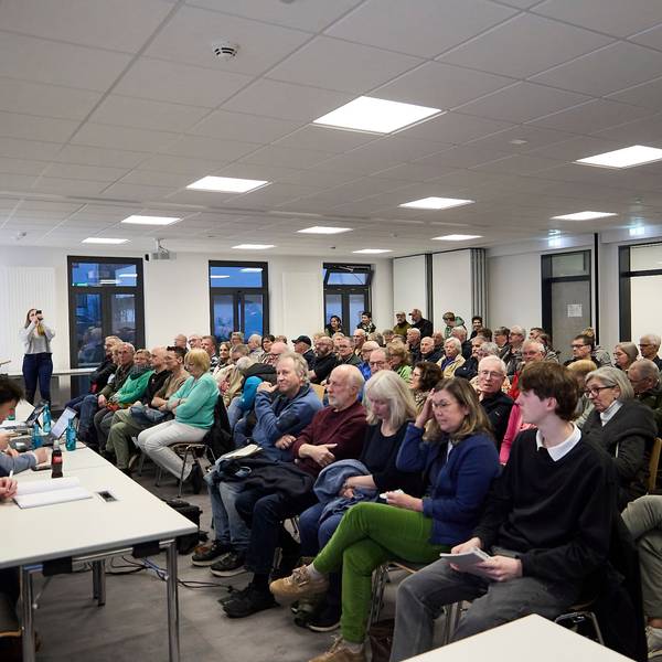 Das Foto zeigt den Versammlungsraum, in dem das Publikum in Reihen sitzt.