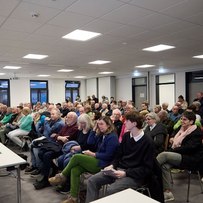 Das Foto zeigt den Versammlungsraum, in dem das Publikum in Reihen sitzt.