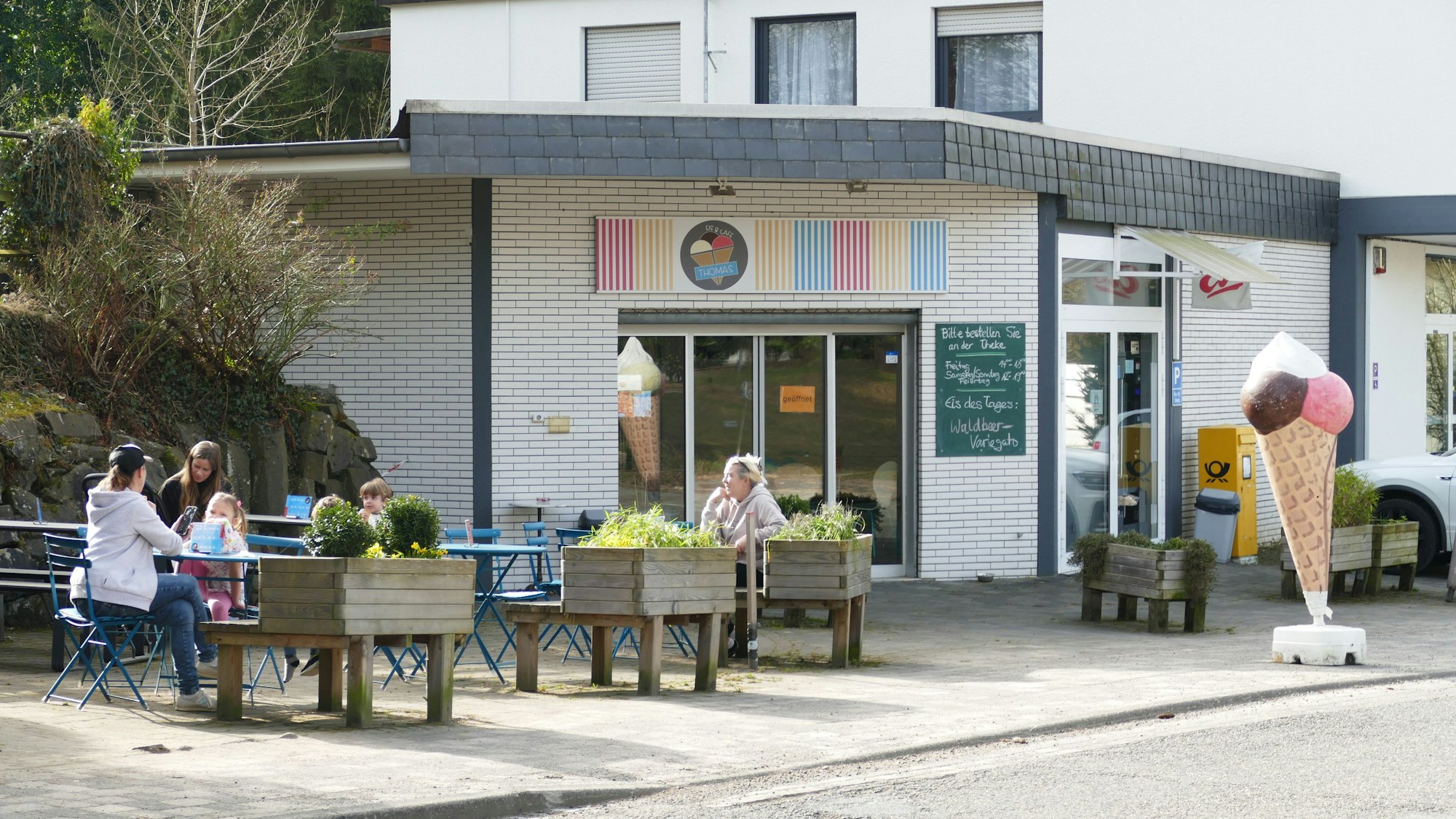 Außenansicht des Eiscafés. Menschen sitzen draußen in der Sonne und vor der Tür steht eine große Eistüte.