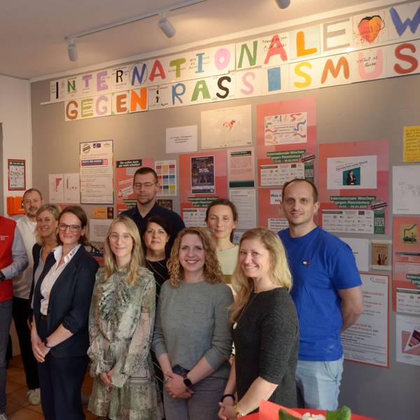 Die Gruppe der oben genannten Vertreter und Vertreterinnen sten vor eine Wand, die mit Plakten gegen Rassismus versehen ist.
