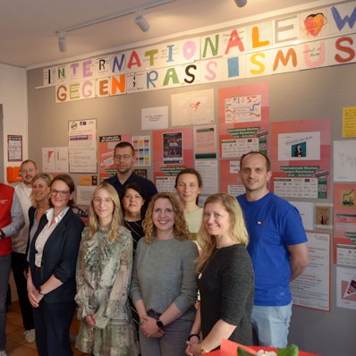 Die Gruppe der oben genannten Vertreter und Vertreterinnen sten vor eine Wand, die mit Plakten gegen Rassismus versehen ist.