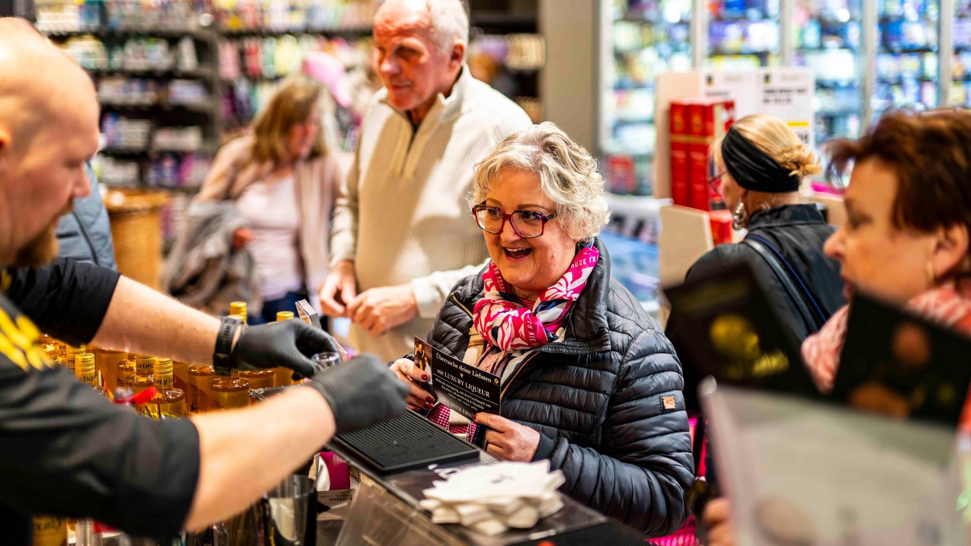 Das Bild zeigt die Seniorin während des Events. Sie steht an einer Theke und wartet auf einen Liquör.