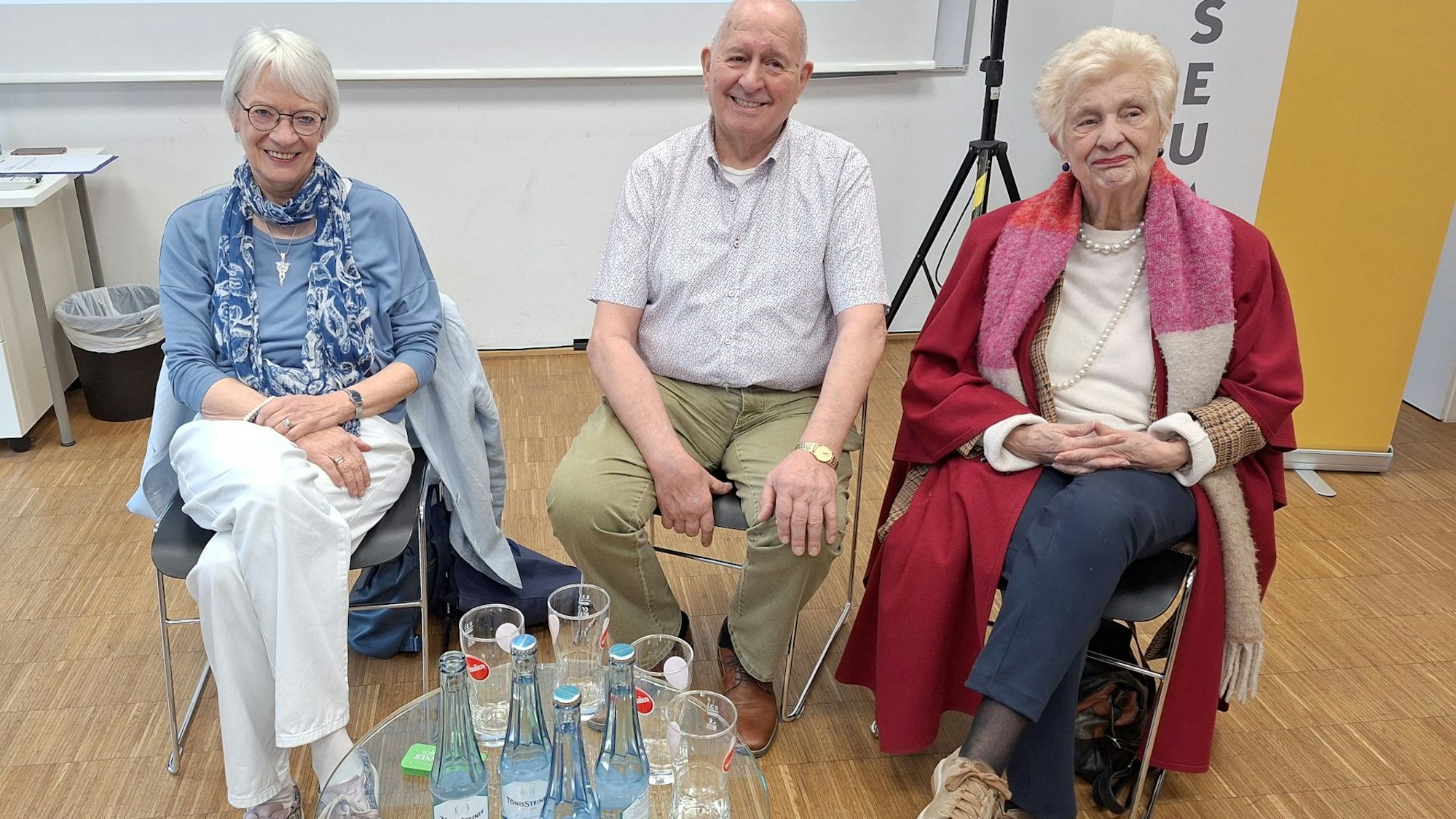 Die drei Obengenannten sitzen auf Stühlen hinter einem Glastisch.