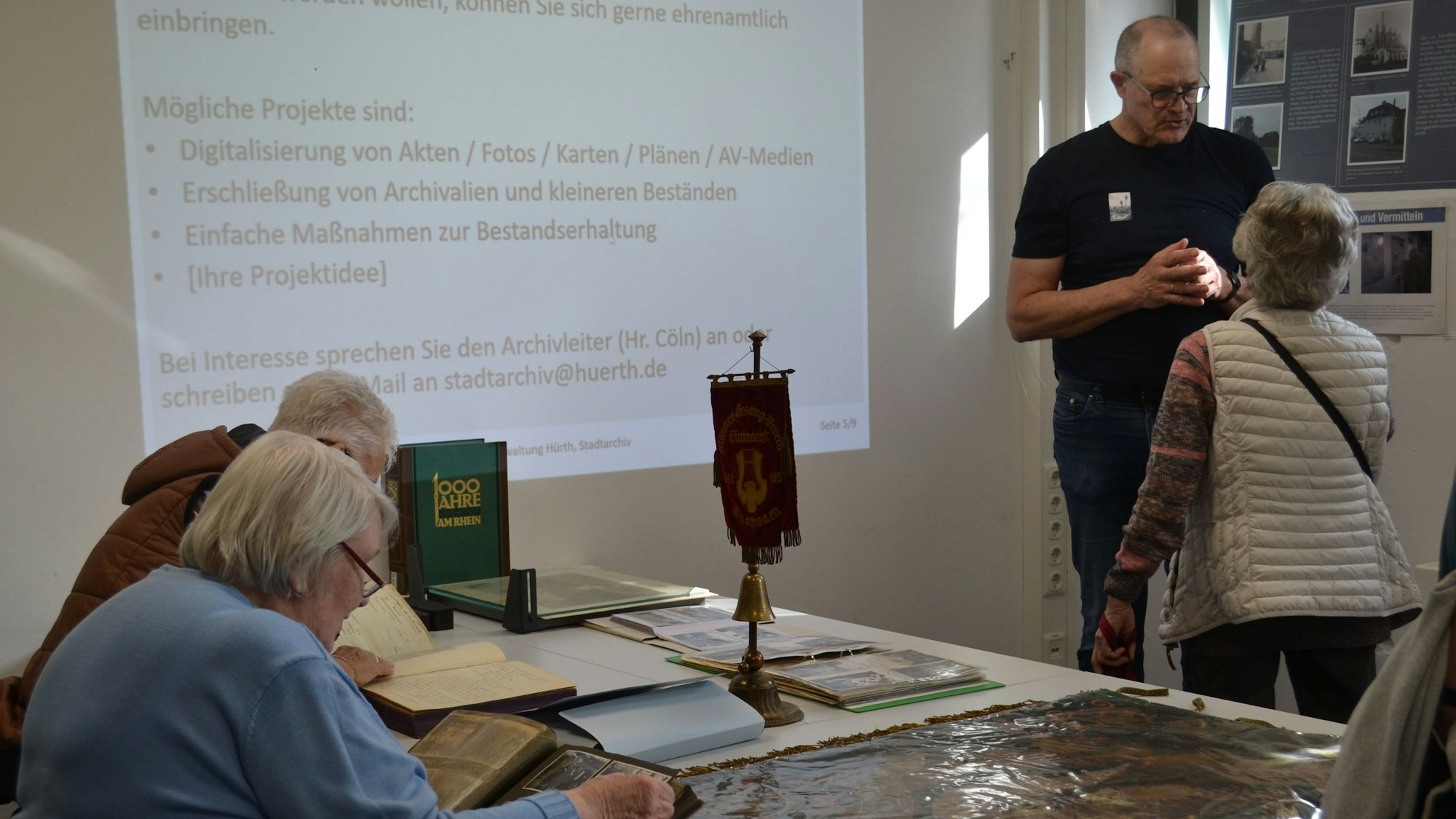 Ein Mann ist in ein Gespräch mit einer Frau vertieft, im Vordergrund blättern Besucher des Archivs in historischen Dokumenten.