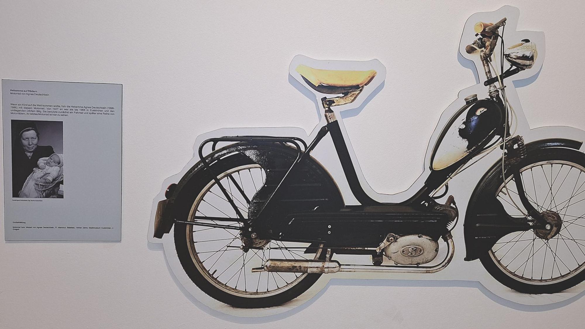 Neben einem großen Ausschnitt eines Fahrrads hängt ein Blatt mit einem Bild der Hebamme Agnes Deutschbein und einem Text an einer Wand.