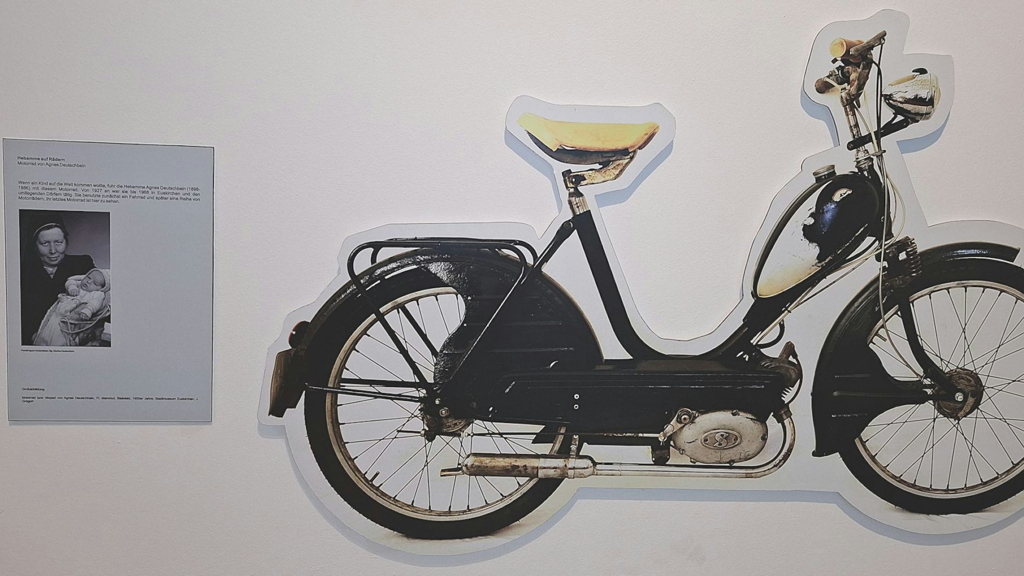 Neben einem großen Ausschnitt eines Fahrrads hängt ein Blatt mit einem Bild der Hebamme Agnes Deutschbein und einem Text an einer Wand.