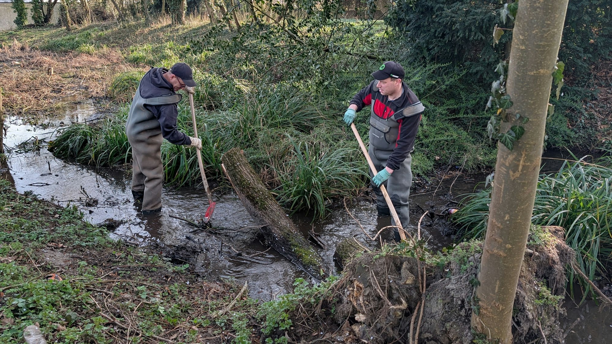 Das Foto zeigt zwei Bauarbeiter in Wasserschutzhosen und mit Spaten im Bachlauf des Gleueler Bachs.