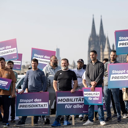 Teilnehmer einer Demonstration halten Schilder mit der Aufschrift „Stoppt das Preisdiktat“ in den Händen. Die Kundgebung richtet sich gegen Pläne der Stadt Köln zur Einführung von Mindestpreisen bei Unternehmen wie Uber und Bolt.