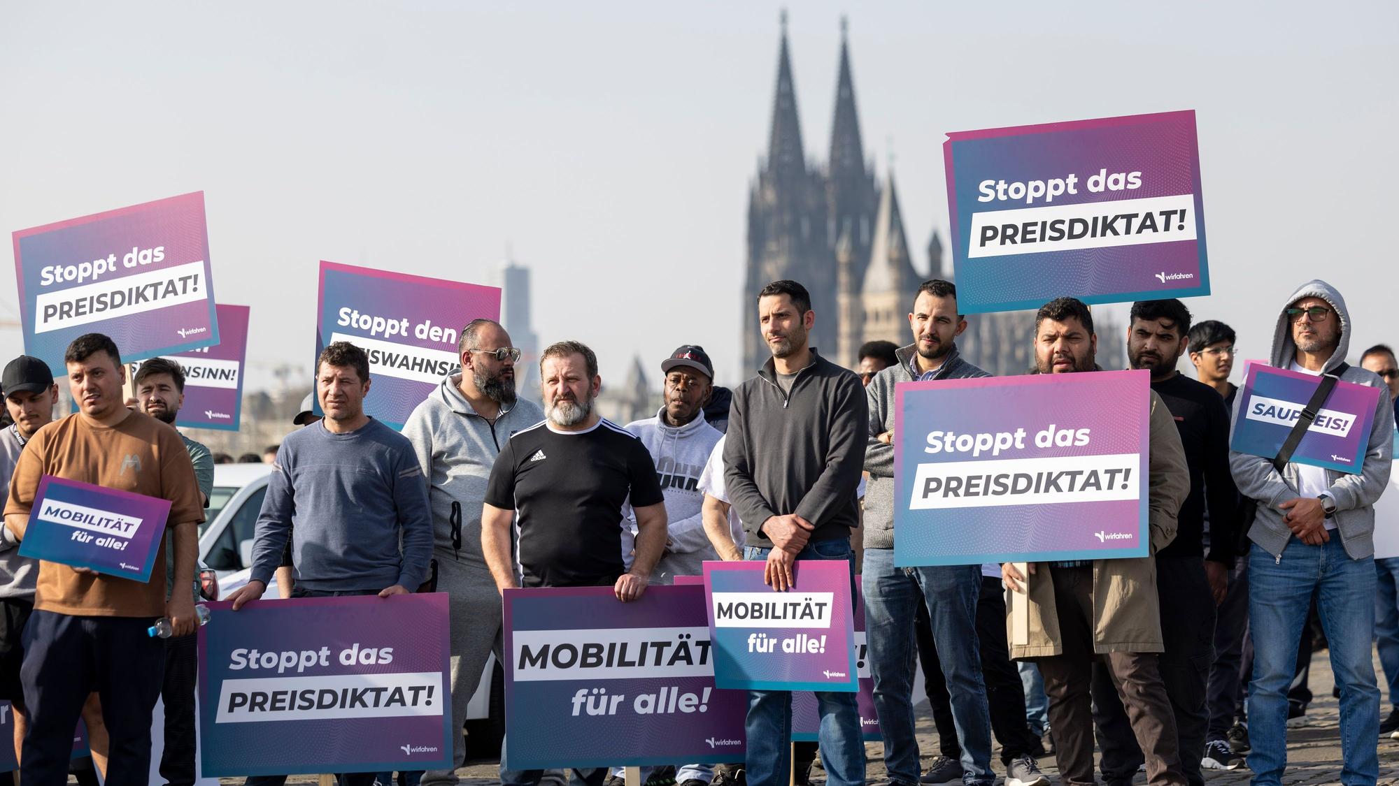 Teilnehmer einer Demonstration halten Schilder mit der Aufschrift „Stoppt das Preisdiktat“ in den Händen. Die Kundgebung richtet sich gegen Pläne der Stadt Köln zur Einführung von Mindestpreisen bei Unternehmen wie Uber und Bolt.