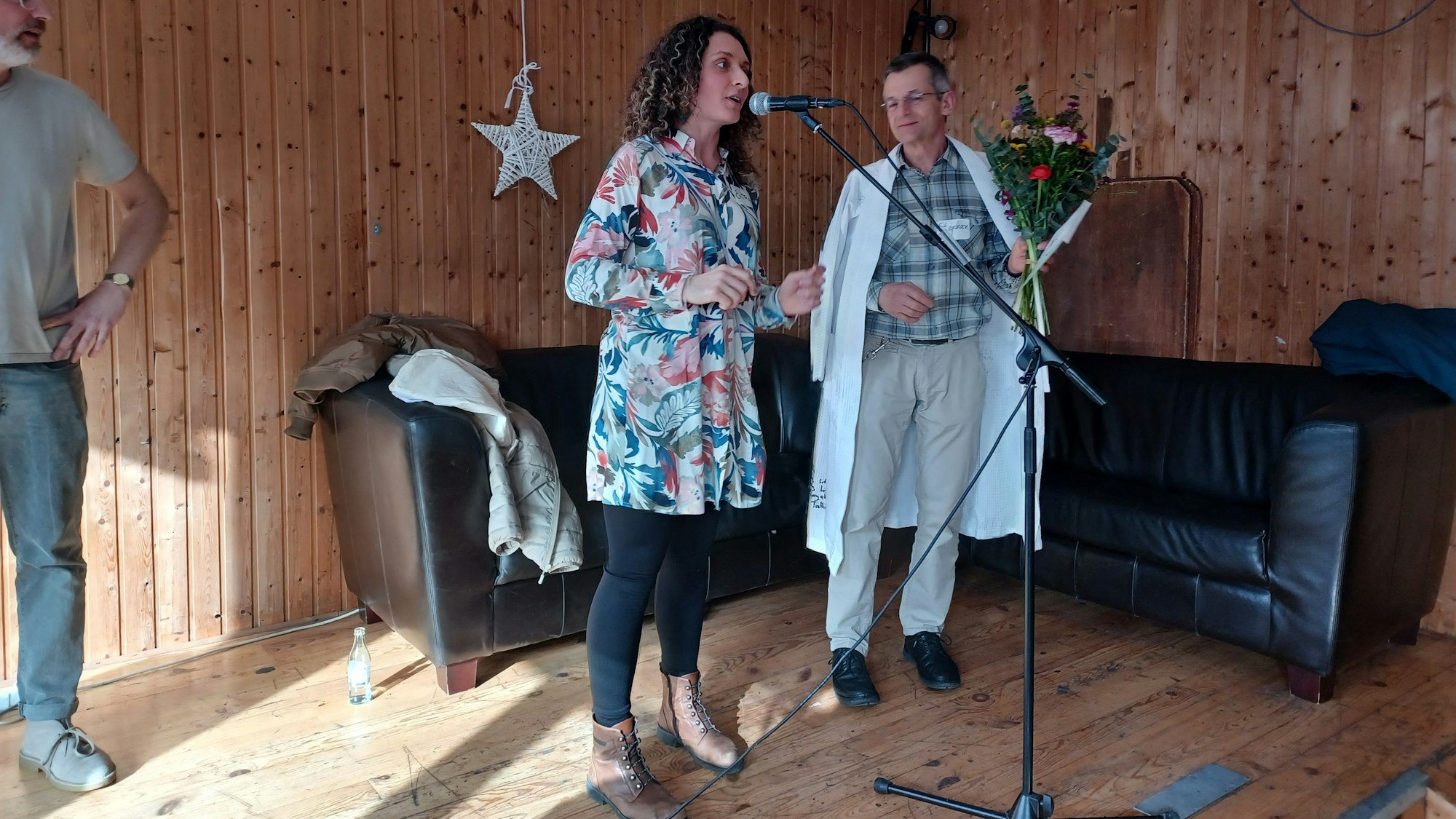 Eine Frau und ein Mann mit Bademantel und Blumenstrauß stehen vor einem Ecksofa.