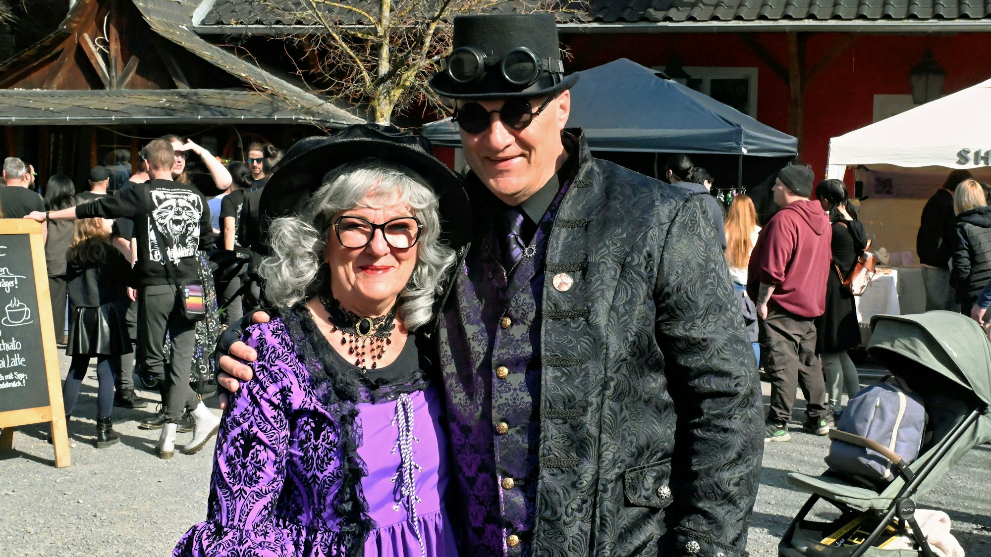 Die oben genannten Eheleute sind in dunklen Kostümen verkleidet und stehen inmitten von Marktbuden.