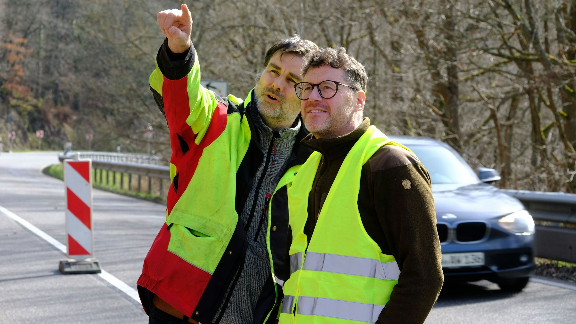 Florian Krumpen und Tobias Wiesen tragen gelbe Warnwesten und schauen den Fällarbeiten zu. Krumpen deutet mit dem ausgestreckten Arm in Blickrichtung der beiden Männer.