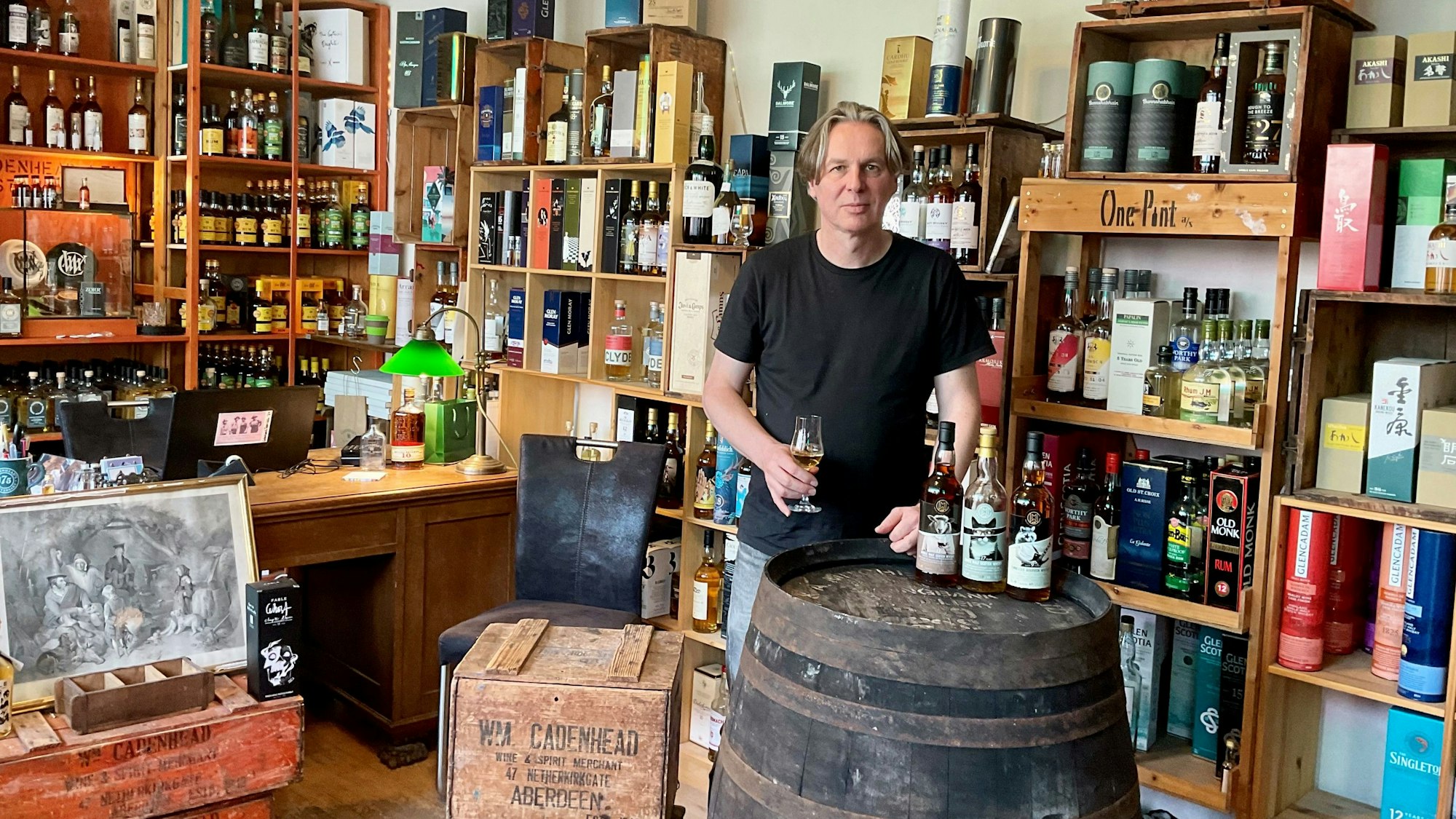 Ein Mann im schwarzen T-Shirt steht in seinem Whiskyladen neben einem Whiskyfass, auf dem Flaschen stehen. Er hält ein Glas in der Hand.
