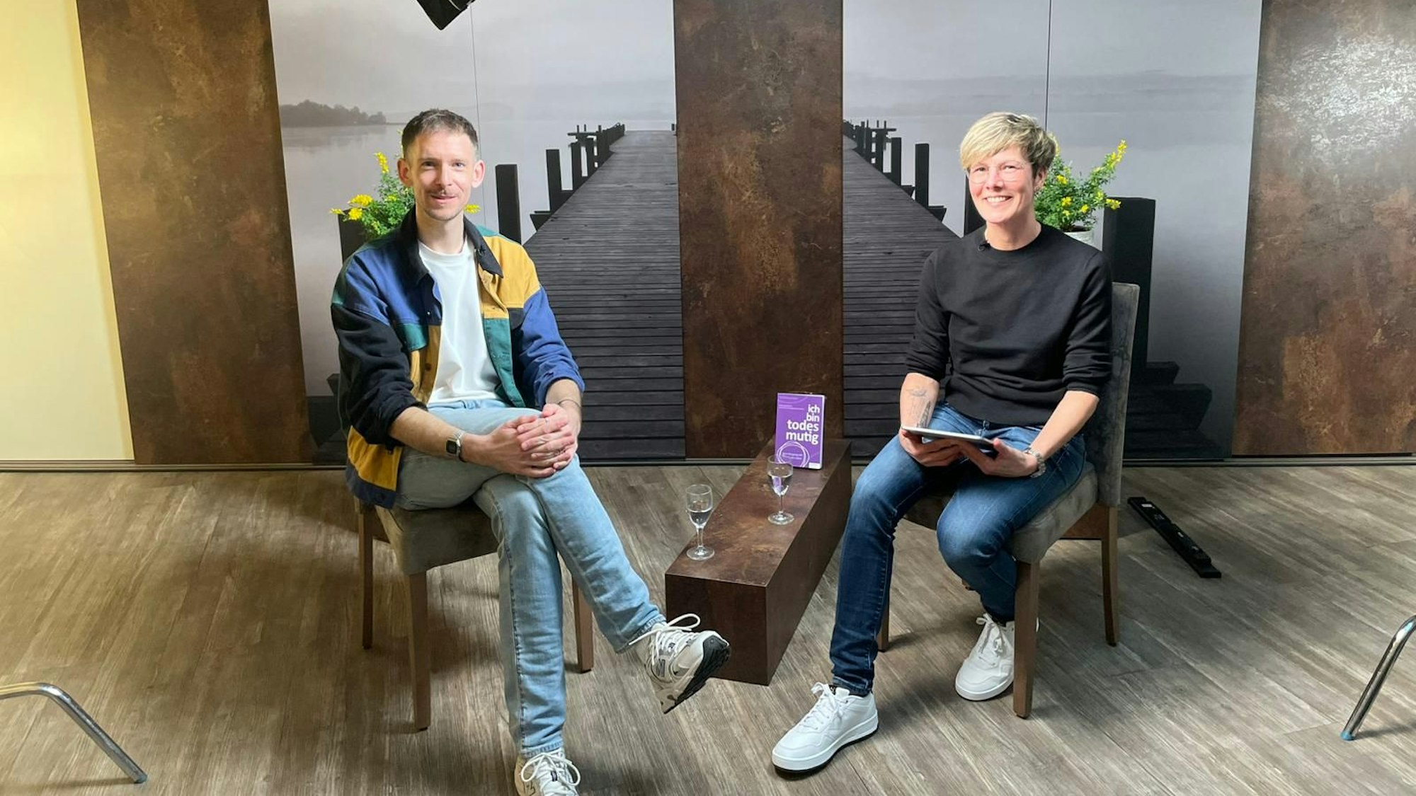 Ein Mann und eine Frau sitzen vor einer Leinwand und schauen in die Kamera.