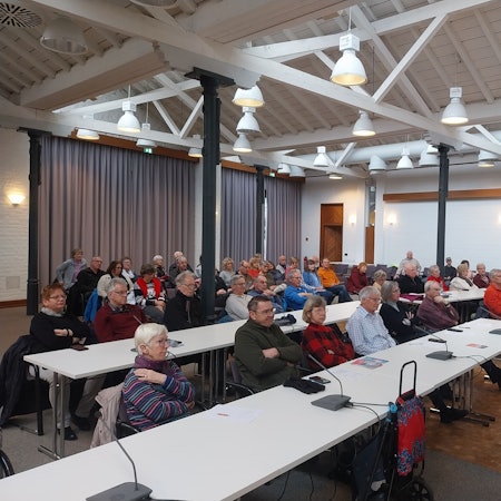 Menschen sitzen an weißen Tischen und blicken aufmerksam nach vorne.