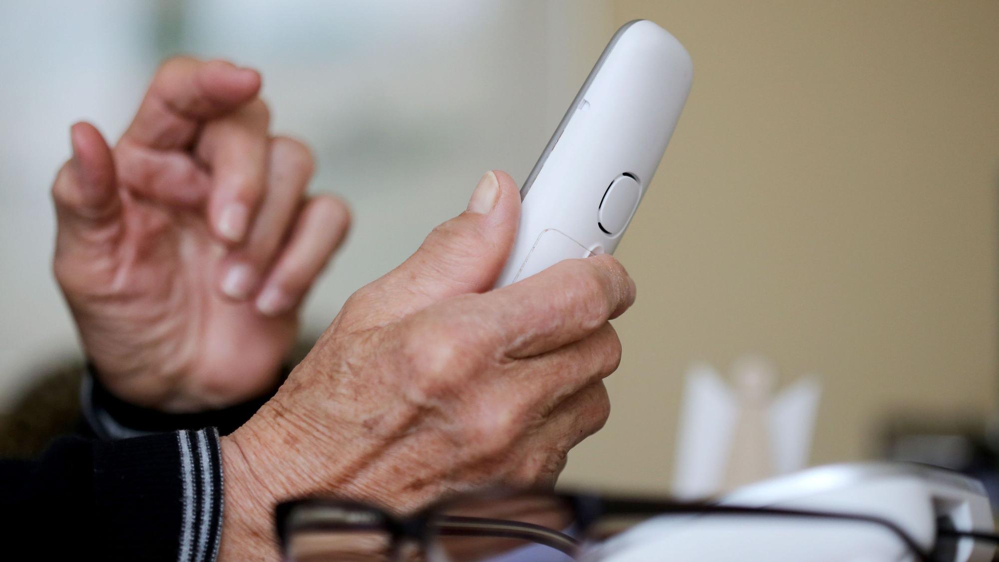 Das Foto zeigt Hände einer älteren Person. In einer Hand befindet sich ein Telefon.