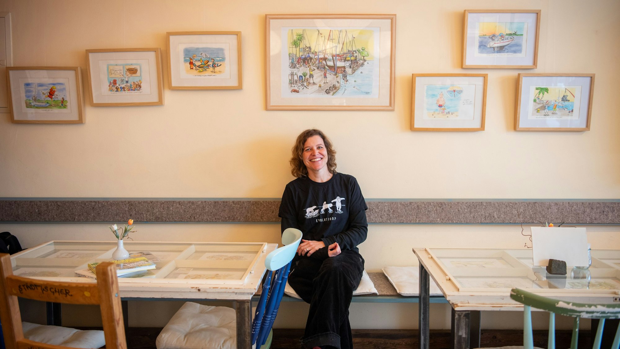 Inga Beitz-Svechtarov sitzt im Fair-Café in Zülpich vor einer Wand, an der ihre Cartoons hängen. Im Vordergrund sind Tische und Stühle zu erkennen.