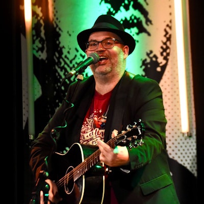 Eni Mann mit Hut und Gitarre auf einer KOnzertbühne.