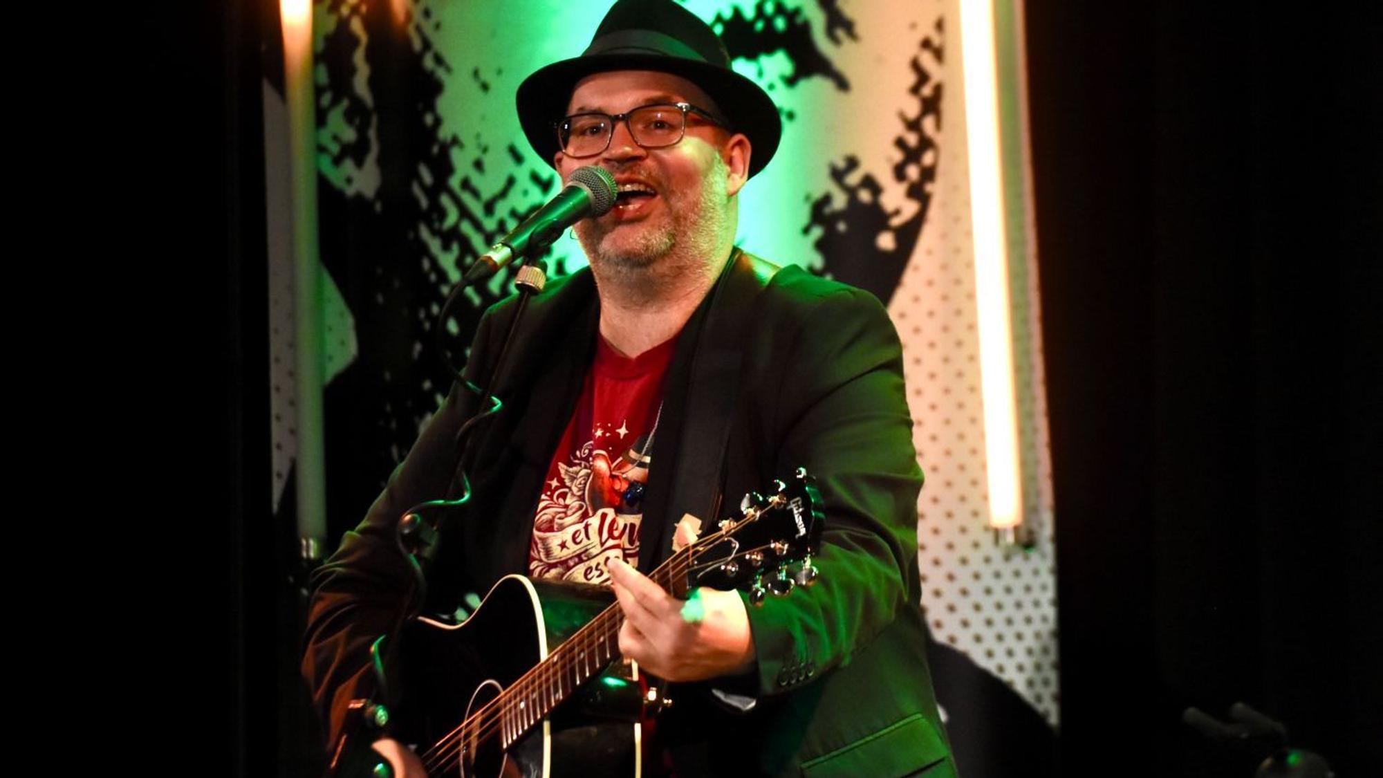 Eni Mann mit Hut und Gitarre auf einer KOnzertbühne.