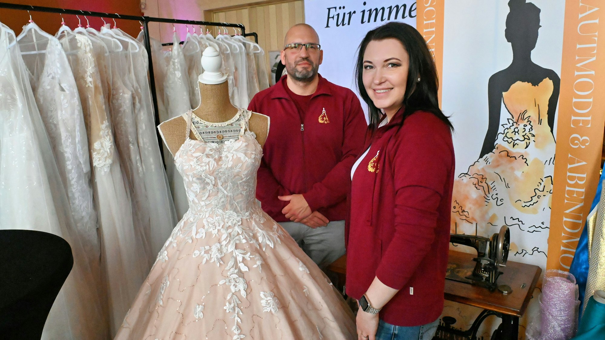 Die beiden Anbieter in roten Jacken stehen neben einer Modepuppe mit einem Hochzeitskleid.