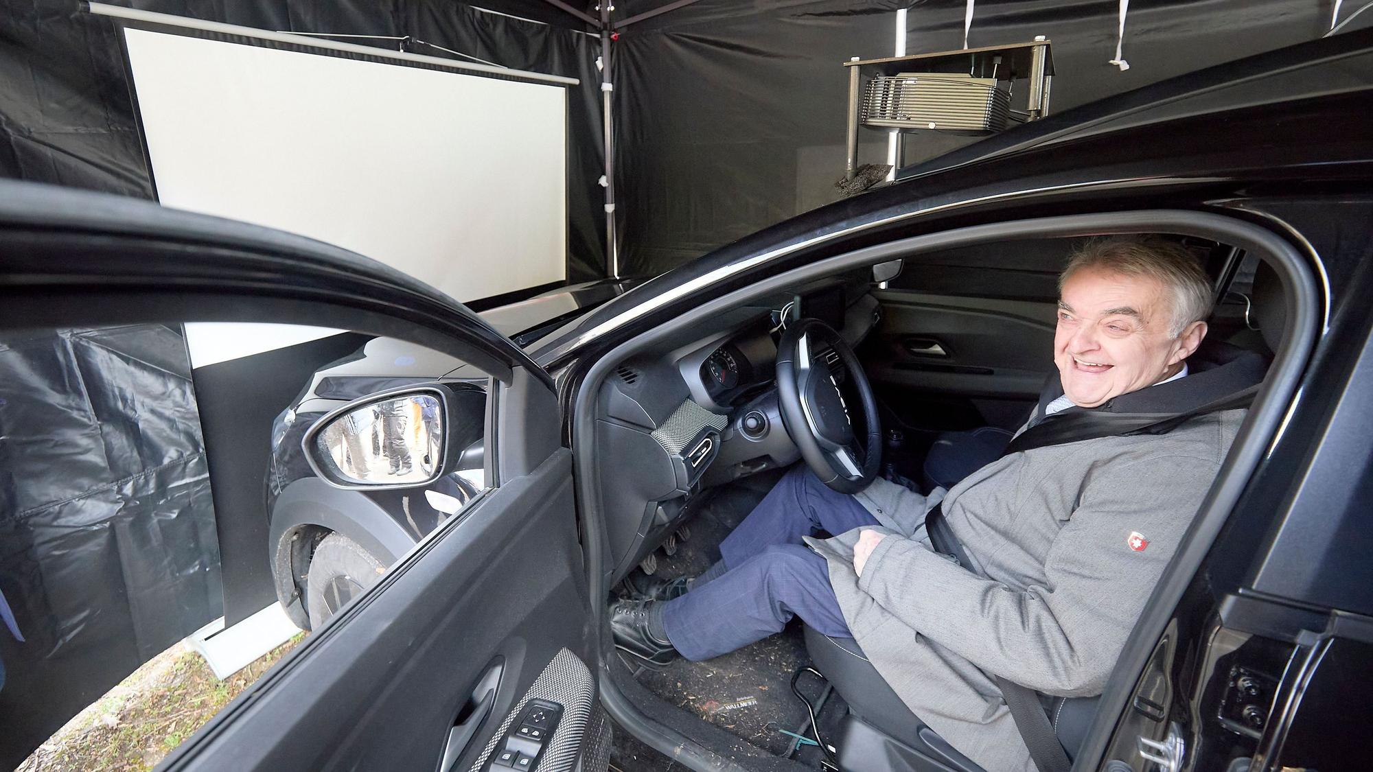 Herbert Reul am Steuer eines Autos, das vor der Leinwand eines Alkoholsimulators steht.