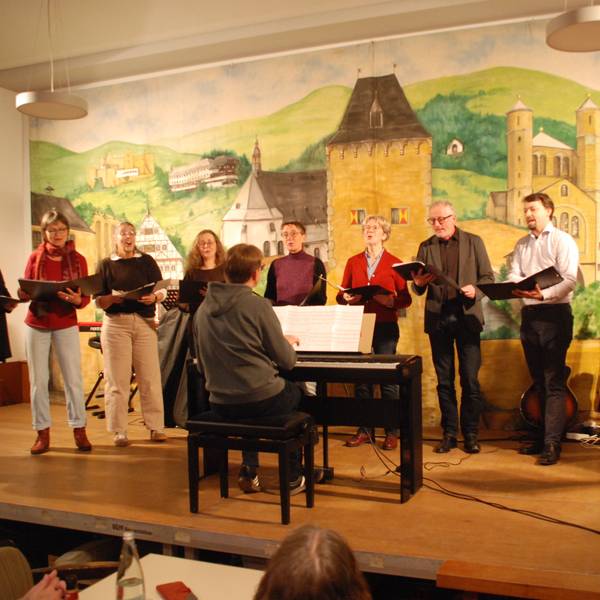 Die Sängerinnen und Sänger stehen in einer Reihe auf der Bühne. Der Dirigent sitzt vor ihnen an einem E-Piano.