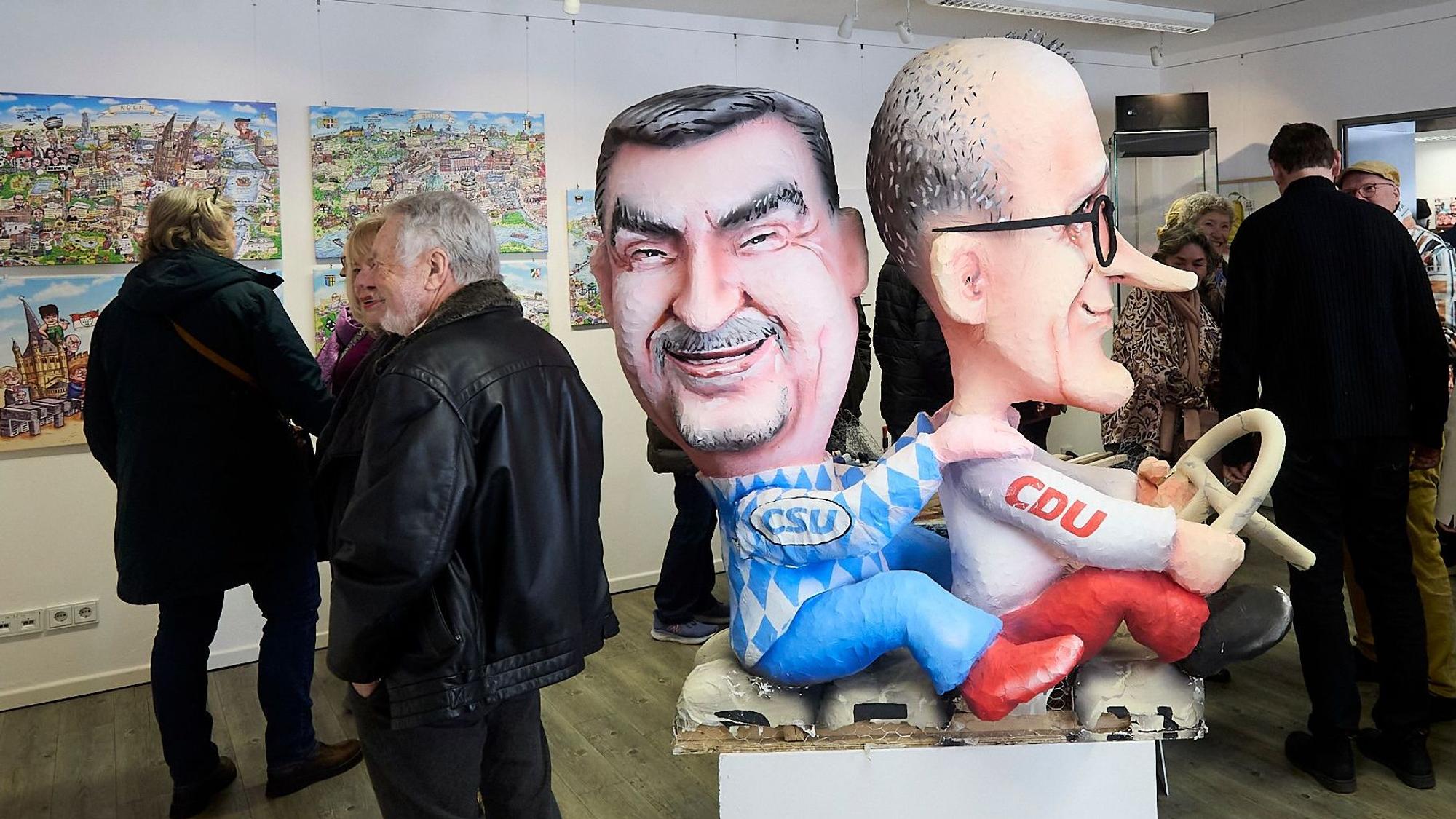 Die Figuren von Markus Söder und Friedrich Merz sitzen hintereinander, Merz hält ein Lenkrad. Im Hintergrund betrachten Menschen die Exponate.