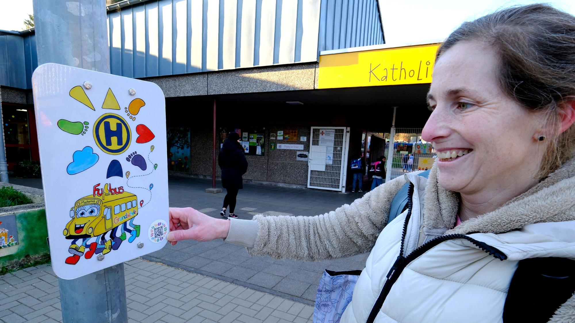 Eine Frau steht an der Grundschule in Mechernich an einem Schild, das die Haltestelle des Fußbusses kennzeichnet.