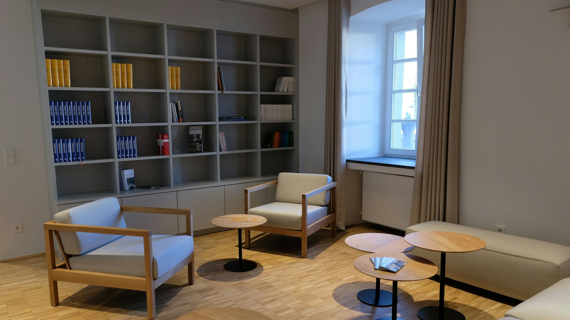 Ein Gemeinschaftsraum mit hellem Parkett und hellen Sitzmöbeln. An der Wand eine Regalwand mit geistlichen Büchern.