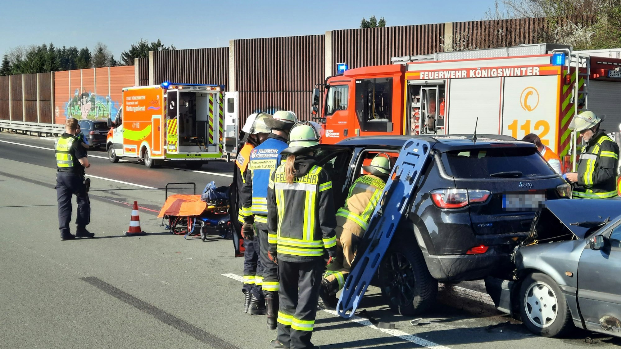 Rettungskräfte an zwei kollidierten Fahrzeugen, im Hintergrund ein Rettungswagen und ein Feuerwehrfahrzeug.
