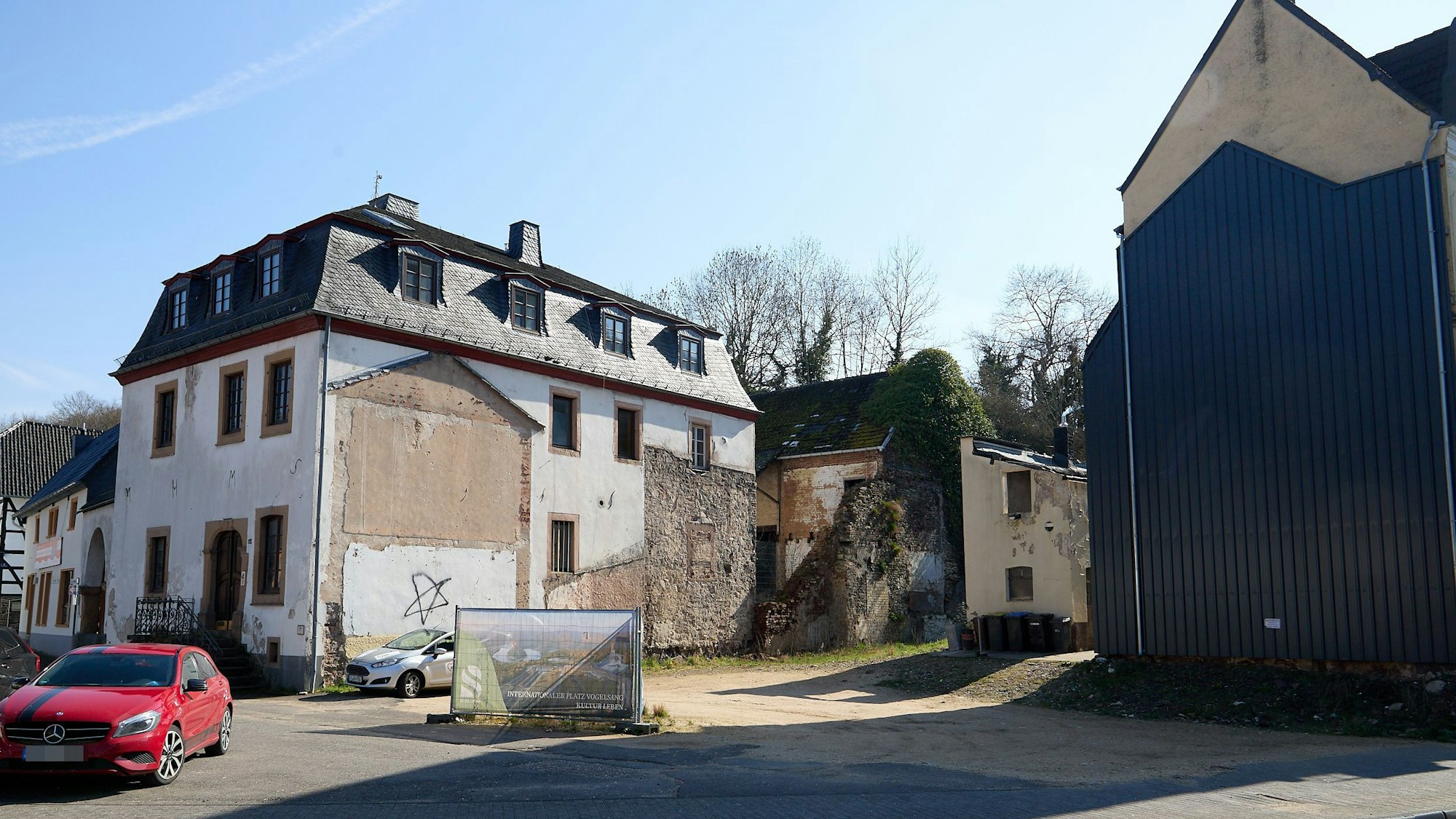 Ein Haus zeigt deutliche Schäden, daneben liegt ein Grundstück brach. Links im vordergrund steht ein rotes Auto.