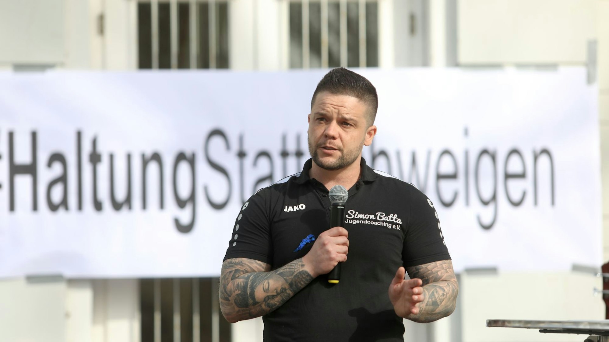 Ein Mann mit Mikrofon, im Hintergrund ein Transparent „Haltung statt Schweigen“.