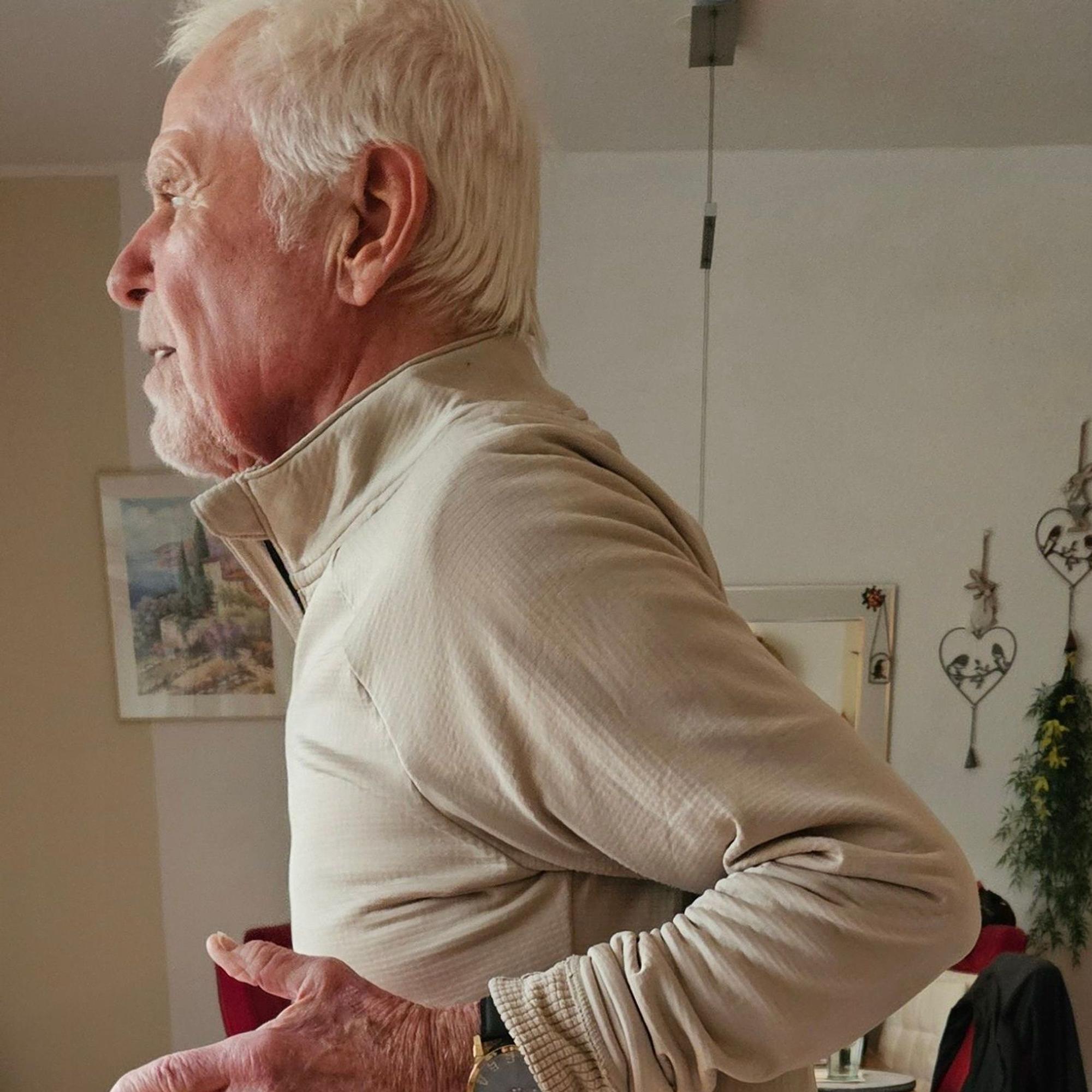 Ein Senior im Gymnastikdress führt die Ellenbogen hinter dem Rücken zusammen.