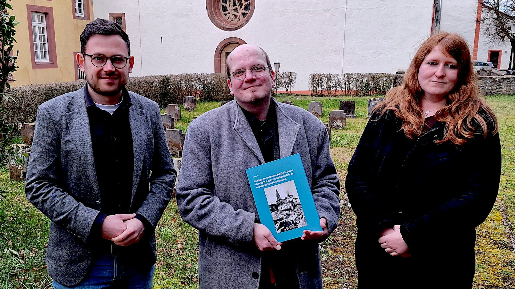 Zwei Männer und eine Frau stehen auf einer Wiese. Im Hintergrund ist ein Teil der weißen Fassade der Basilika in Kall-Steinfeld zu erkennen.