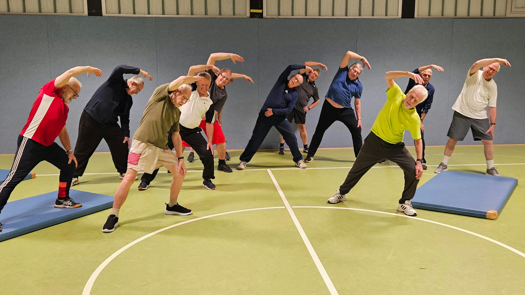 Eine Gruppe von älteren Männern macht Dehnübungen in einer Sporthalle.