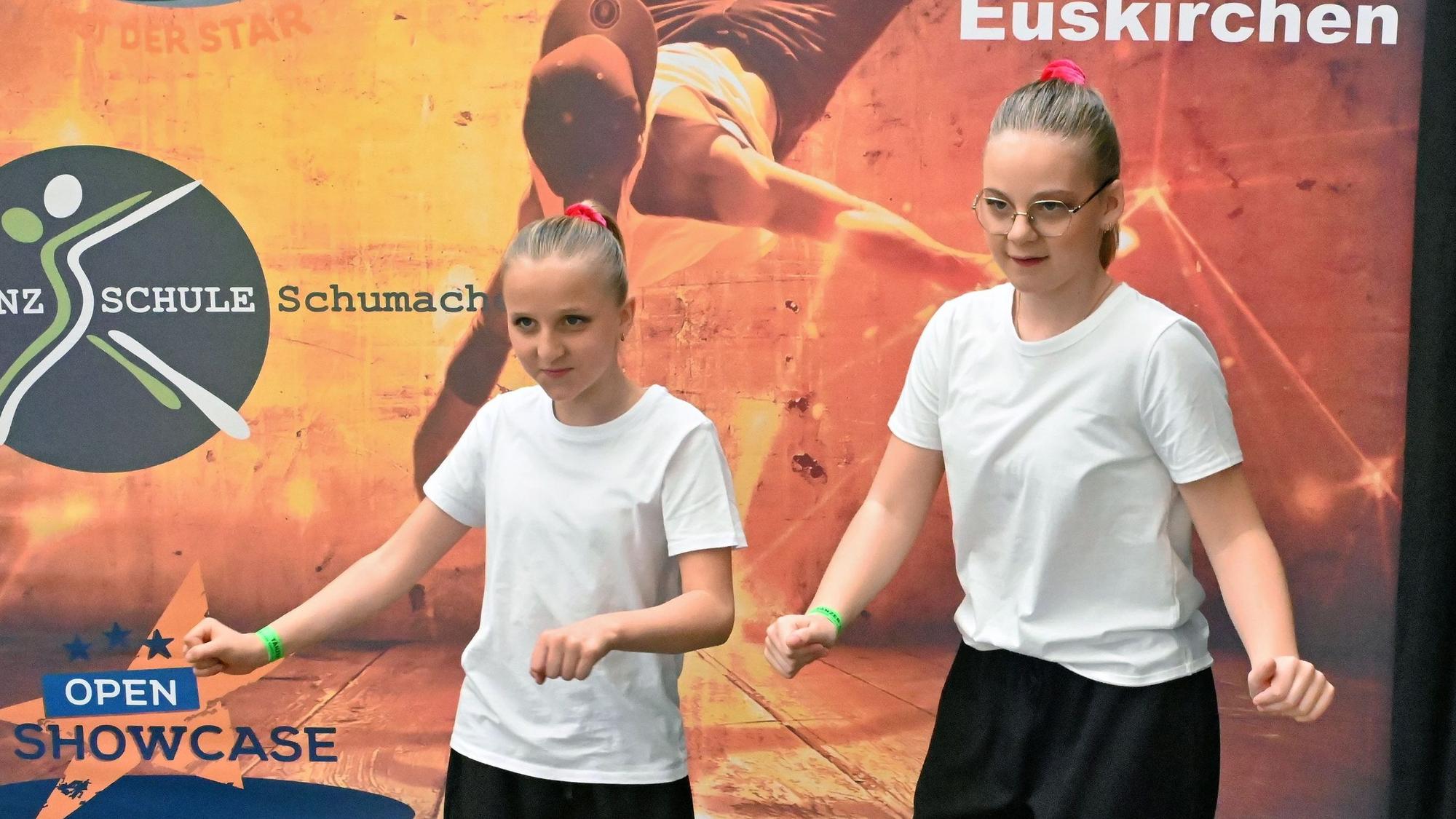 Zwei Mädchen in schwarzen Hosen und weißen Shirts machen Tanzbewegungen.